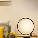 Eine stilvolle 'CircoloLuce' Nachttischlampe in minimalistischem Design leuchtet auf einem Nachttisch, neben einer Vase mit Zweigen.