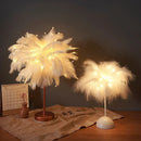 Lampe de chevet 'LuceSoffusa' stylée avec des plumes blanches chaleureusement lumineuses sur un pied en bronze, à côté d'un calendrier, souligné par une télécommande au premier plan.
