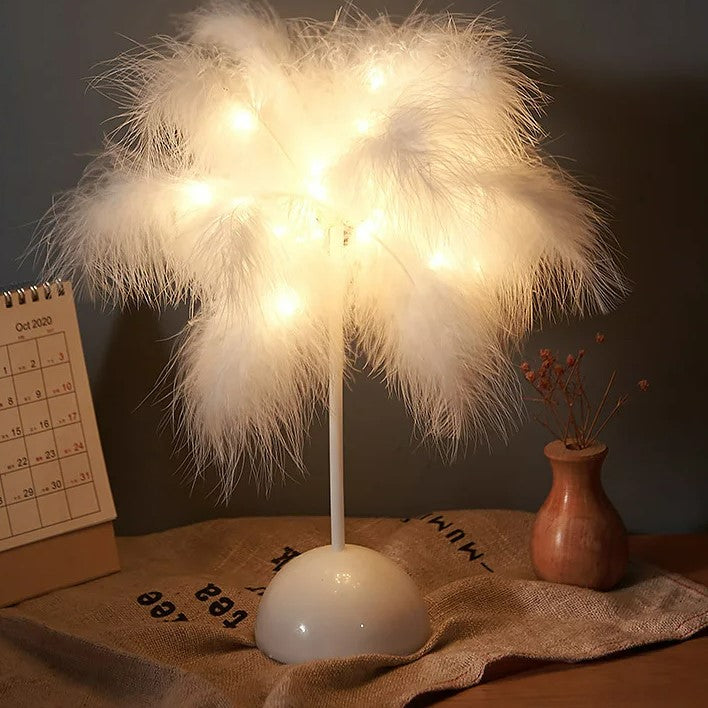 Elegante lampada da comodino "LuceSoffusa" con piume bianche dai riflessi caldi su una base color bronzo, accanto a un calendario, sottolineato da un telecomando in primo piano.